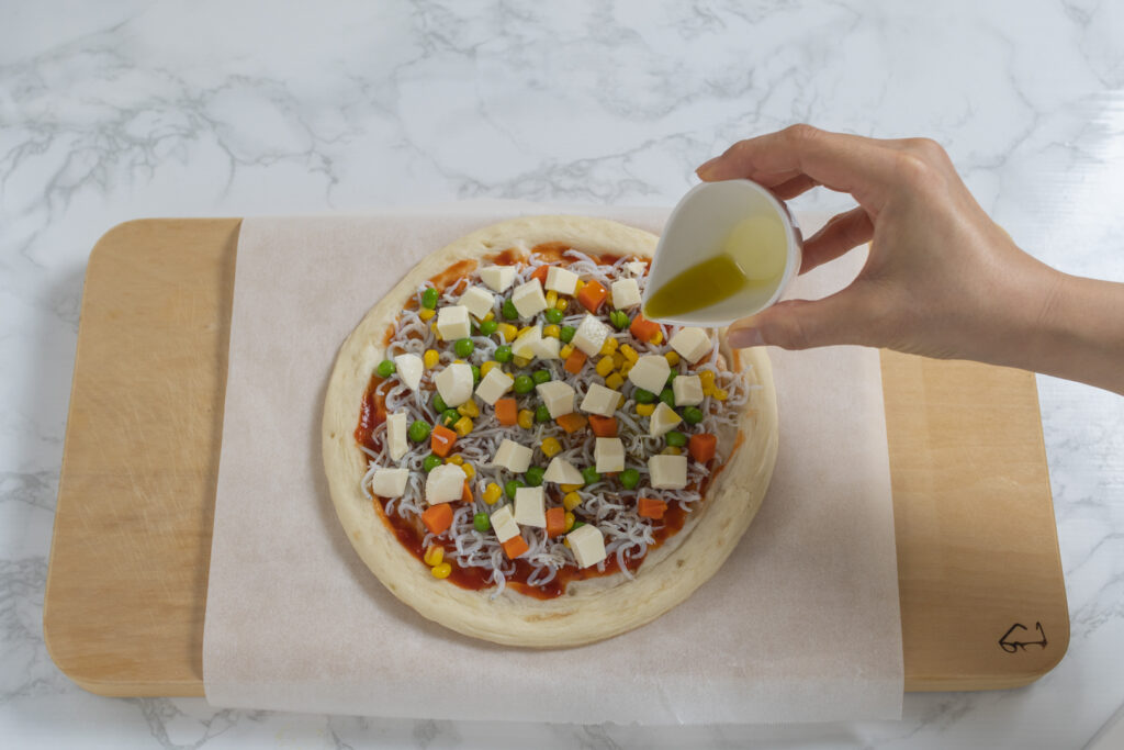 ソースを塗ったピザ生地にしらすとミックスベジタブルとモッツァレラチーズをのせ、オリーブオイルを回しかける。